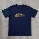 A navy blue short-sleeve crewneck t-shirt with 'LOCAL CELEBRITY' printed in gold across the chest, laid flat on a textured gray concrete surface.